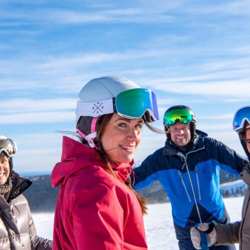 Fire mennesker i skidragter og hjelme står sammen på et snedækket bjerg under blå himmel.