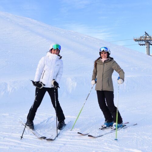 To personer på ski står i sneen med skistave, liftstænger og blå himmel i baggrunden.