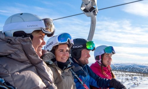 Four people in ski clothes sitting in a chairlift above snowy landscape with blue sky