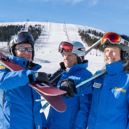 Three people in blue ski jackets stand with skis in front of a snow-covered ski slope and lifts.