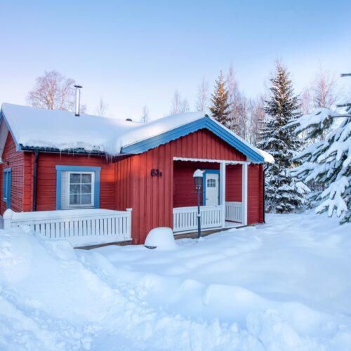 Rød hytte med blå detaljer, snedækket tag, hvid veranda og omgivende grantræer i vinterlandskab.