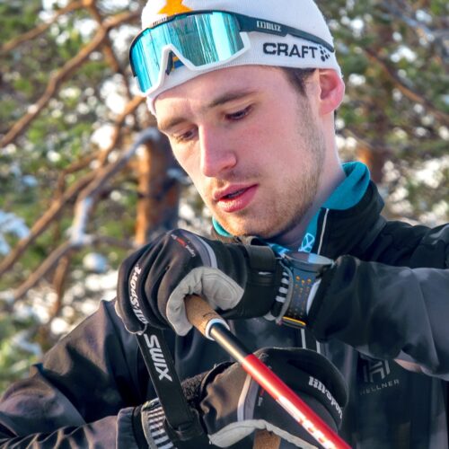 Närbild på person i skidutrustning med svarta handskar, stavar och skidglasögon, skog i bakgrunden