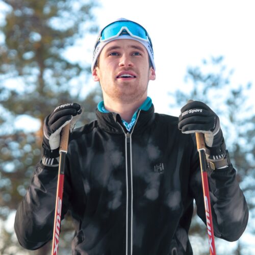 Person i sort skijakke med stave i hånden på snedækket langrendsløjpe med skov i baggrunden