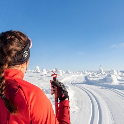 Person i röd skidjacka står vid preparerat längdskidspår i soligt snötäckt fjällandskap