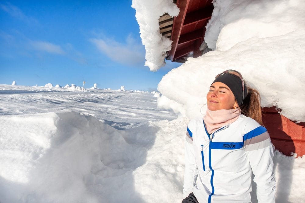 Person in white ski jacket leans against snow-covered cabin in sunny mountain landscape