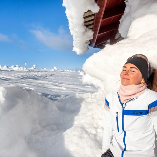 Person i vit skidjacka lutar mot snötäckt stuga i soligt fjällandskap