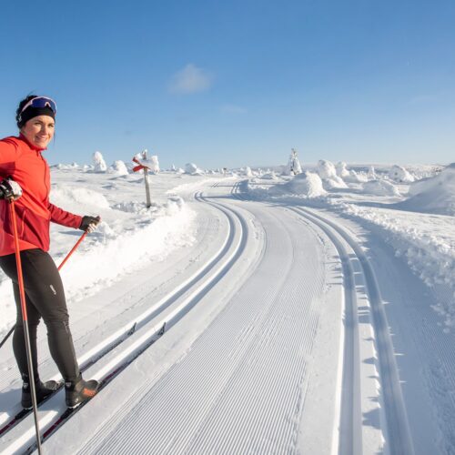 Person i rød skijakke står på præparerede langrendsspor i bjerge med snelandskab og blå himmel