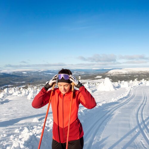 Person i röd skidjacka justerar skidglasögon på preparerade längdskidspår med utsikt över snötäckta fjäll