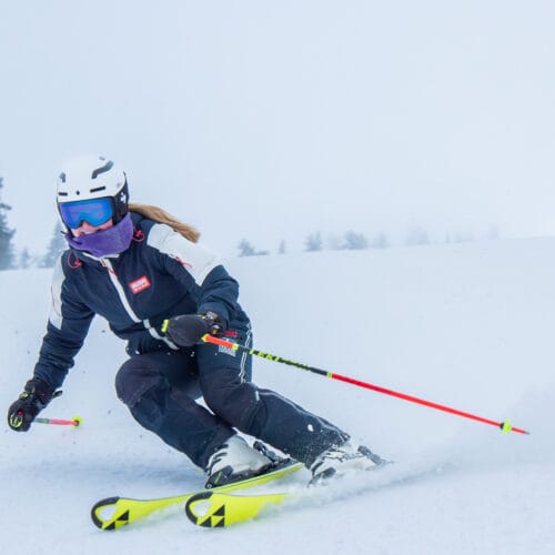 Skidåkare carvingsvänger i öppen pist, vit dimmig fjällmiljö, hjälm, stavar och gula skidor.