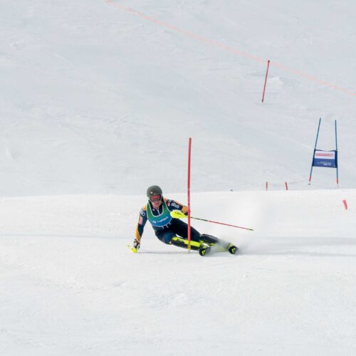 Alpine skier in racing suit makes a sharp turn around red slalom pole on snow-covered slope with several gates in the background