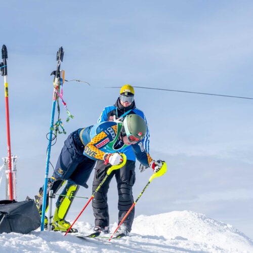 Alpine skiers in racing suits lean forward at the start gate with poles, while equipment and snowy slope are visible in the background