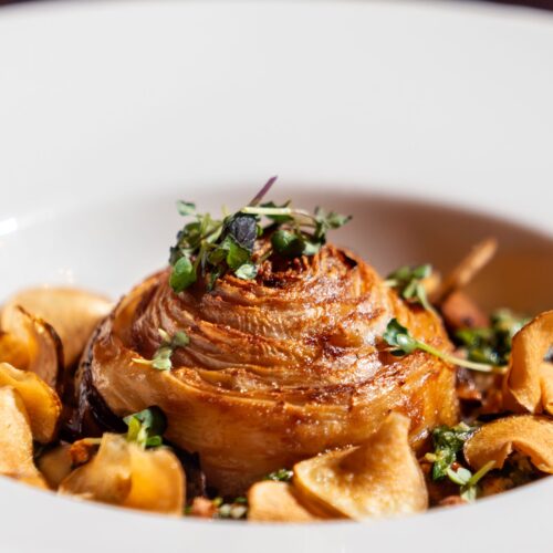 Caramelised potato rose with crispy chips and fresh herbs in deep white plate.