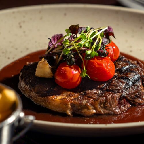 Grilled steak with dark sauce, topped with cherry tomatoes, garlic and fresh herb shoots on light coloured plate.