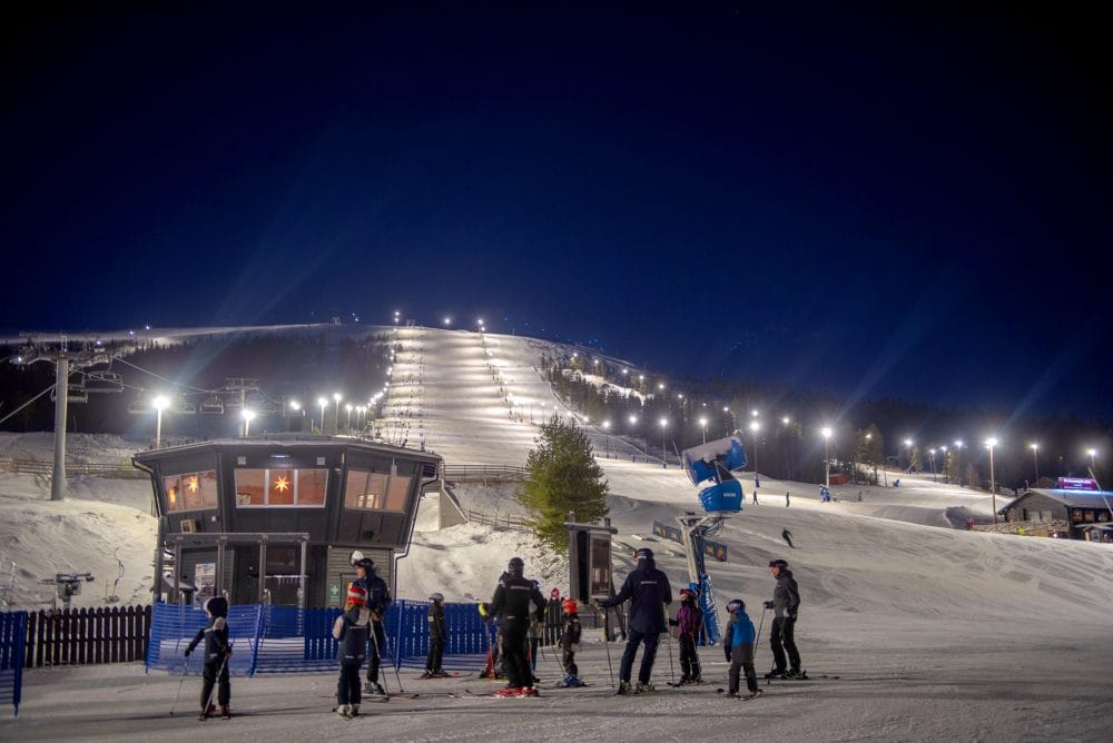 Upplyst skidbacke för kvällsåkning med snökanoner och skidåkare vid foten