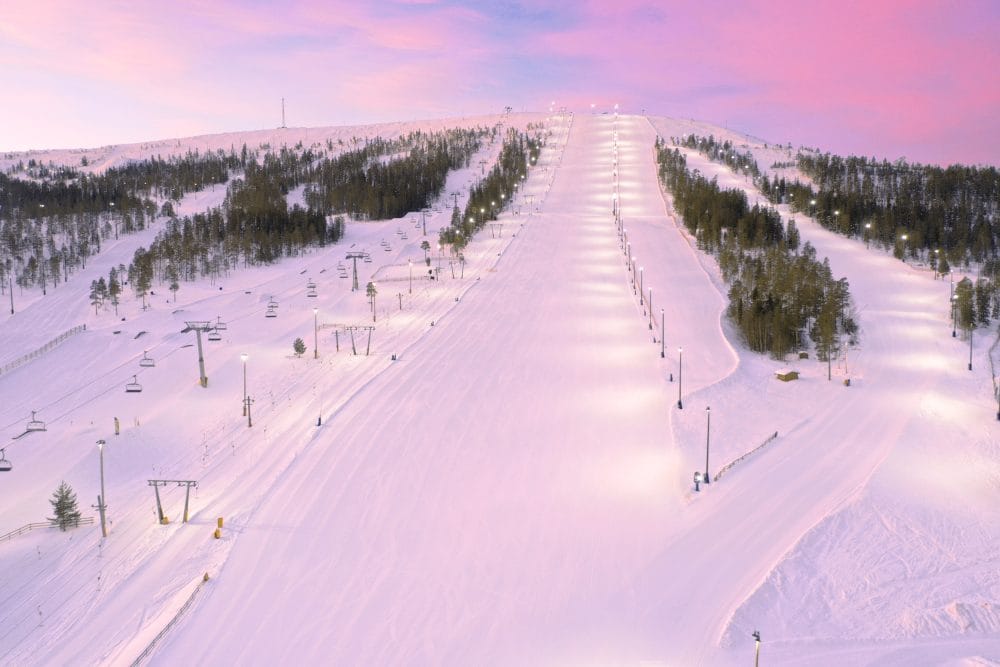 Upplysta skidbackar på fjäll under rosa kvällshimmel med granar längs sluttningen och liftar