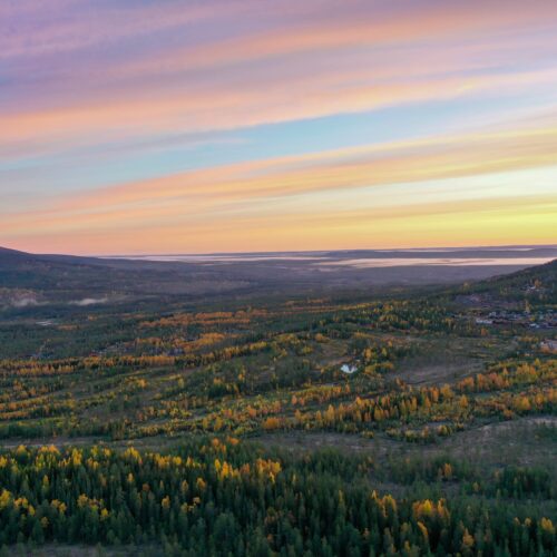 Efterårslandskab med farvede træer og bjerge under bløde pastelfarvede solnedgangsskyer