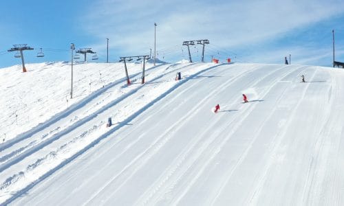 Wide, freshly groomed ski slope with several skiers in red clothes under clear blue skies
