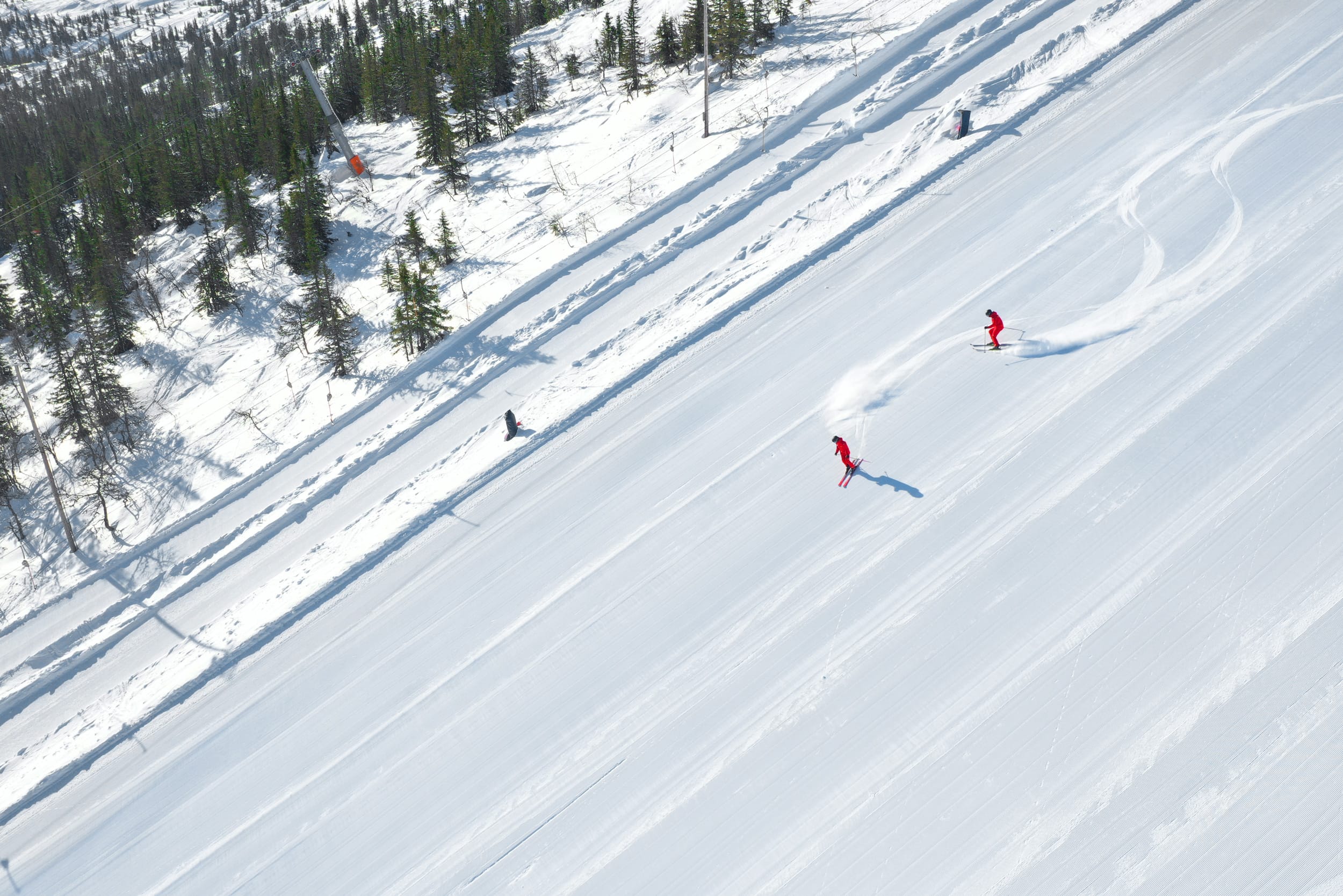 Two skiers in red clothes ski down a wide, freshly groomed ski slope with a pine forest