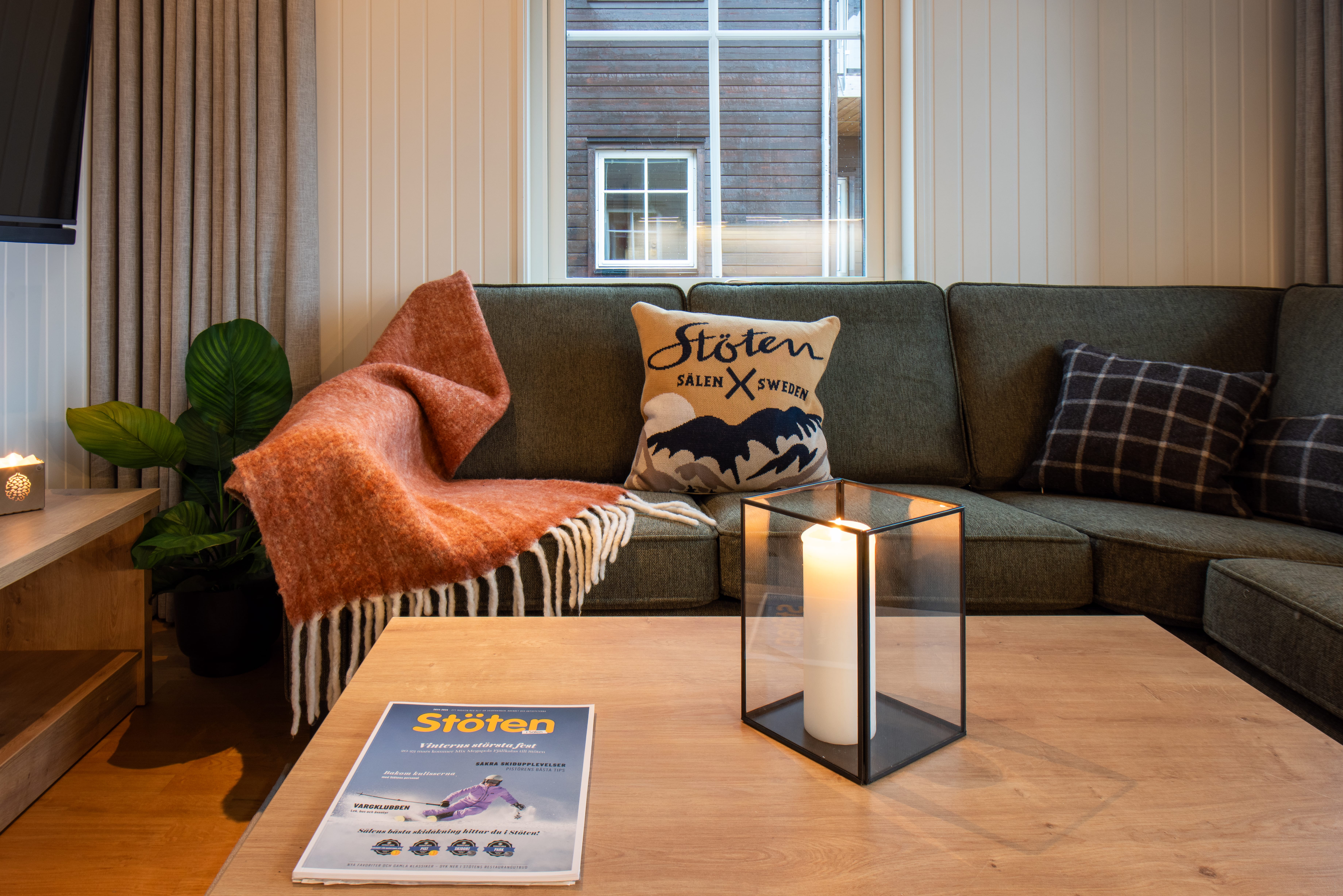 Grön hörnsoffa med kuddar och filt, tänd ljuslykta på trägolv, tidning om Stöten på bordet.