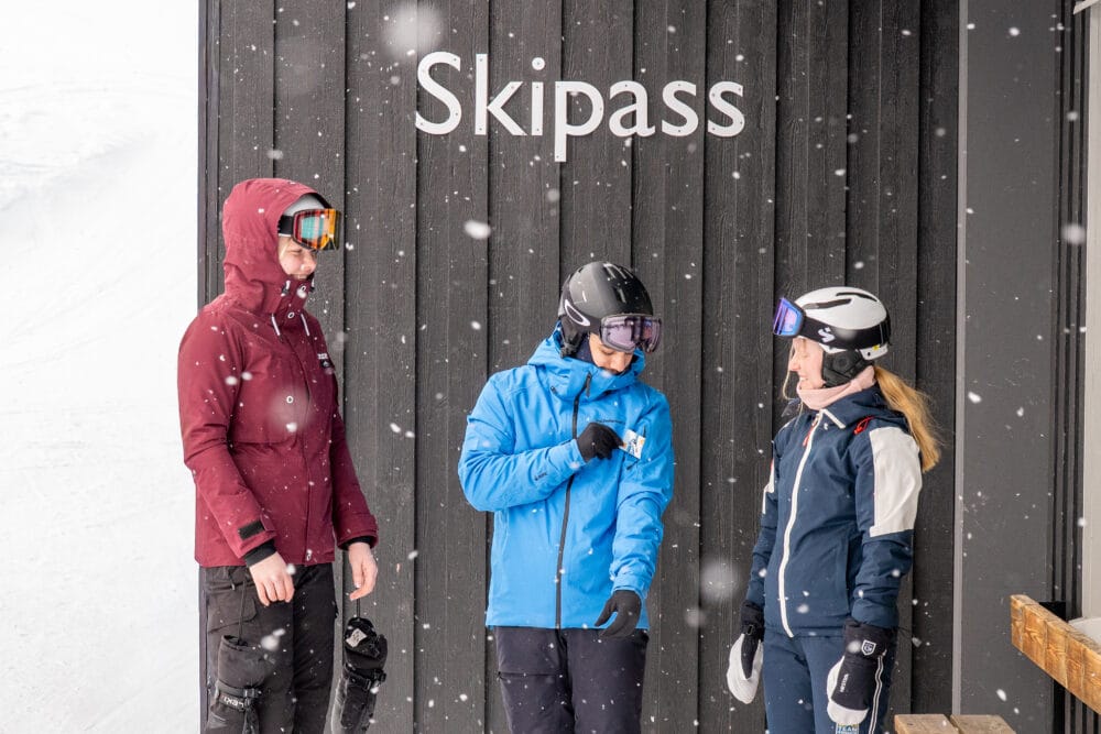 Tre personer i skidkläder står vid skipass-skylt medan snö faller utomhus nära liftområde.