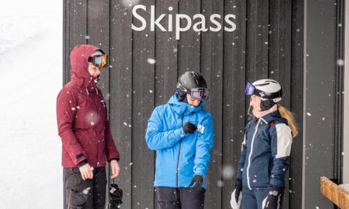 Three people in skiwear stand by ski pass sign while snow falls outside near lift area.
