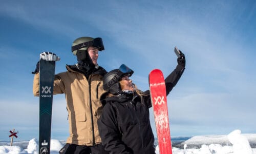 Two people with ski equipment pose with skis in a snowy mountain landscape under a clear sky.