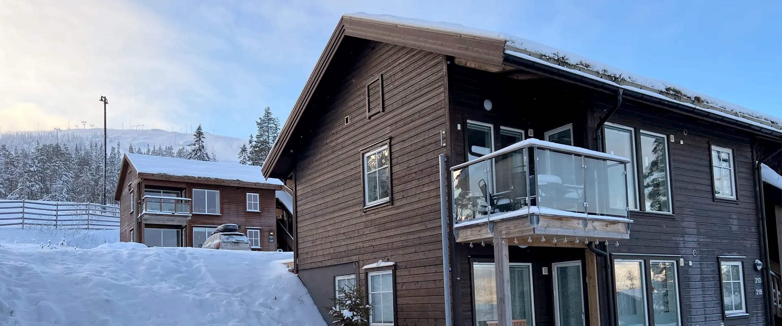 Brunt træhus med glasrækværk på altanen, store vinduer, snedækket jord og baggrund af bjerge og grantræer.