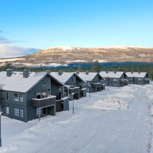 Flere mørkegrå toetagers huse med snedækkede tage, pløjede veje og bjerge i baggrunden under en klar blå himmel.