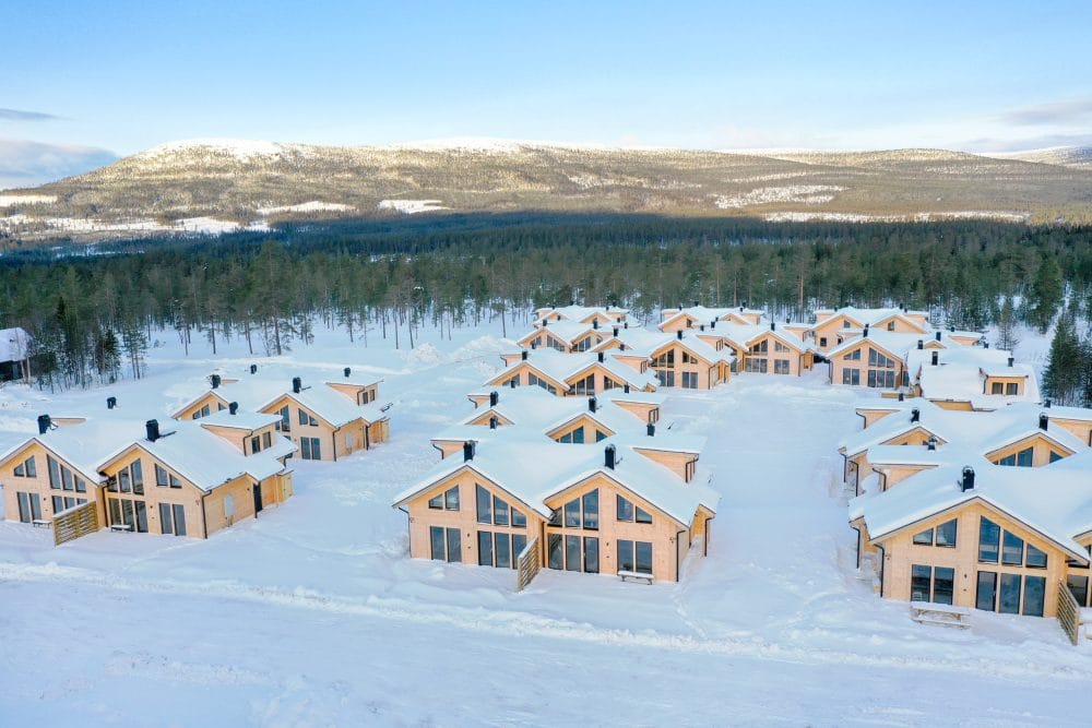 Flera ljusa trästugor med snötäckta tak, omgivande tallskog och fjäll i bakgrunden under vinterdag.