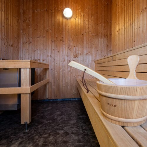 Träpanelklädd bastu med träbänkar, hink och skopa, mörkt golv och rund vägglampa.