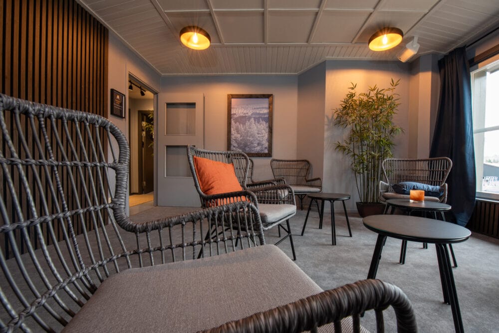 Lounge with rattan chairs and orange cushion, small black tables, green plant in the corner and wall decoration with nature motifs.