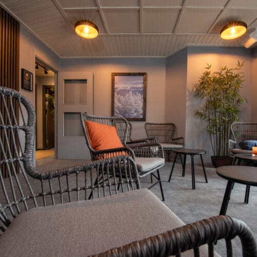 Lounge med rottingstolar och orange kudde, små svarta bord, gröna växten i hörnet och väggdekoration med naturmotiv.