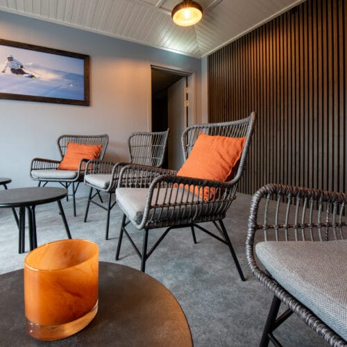 Lounge med rottingstolar och orange kuddar, små svarta bord, vägg med träpanel och skidbild på väggen.