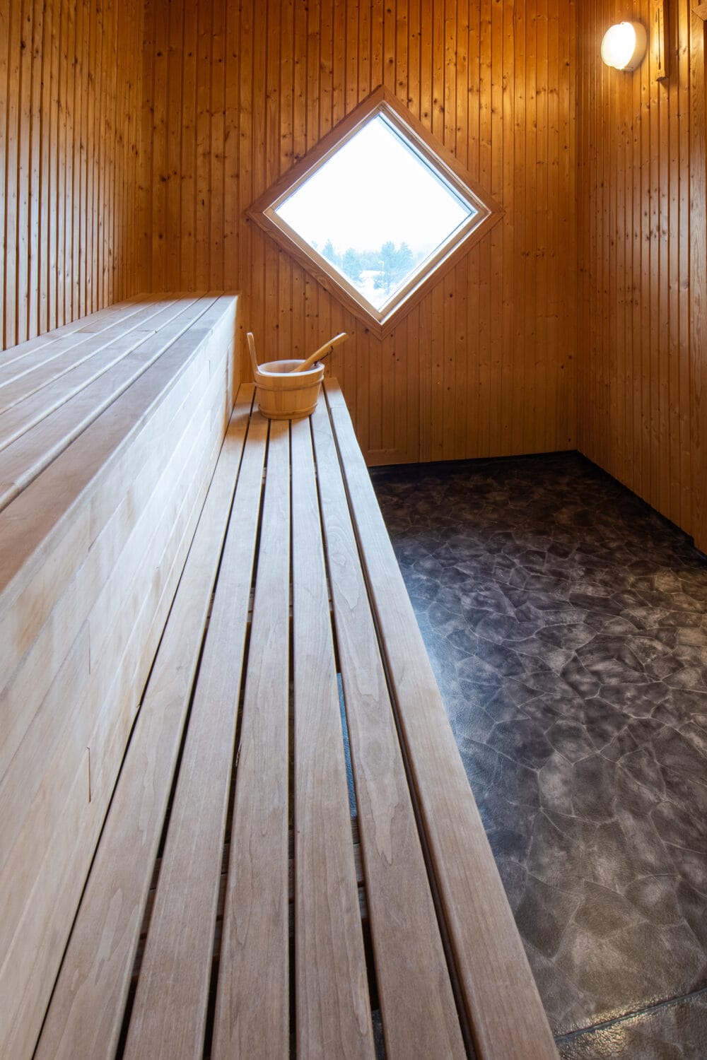 Træbeklædt sauna med træbænke, spand og øse, diamantformet vindue og mørkt gulv.