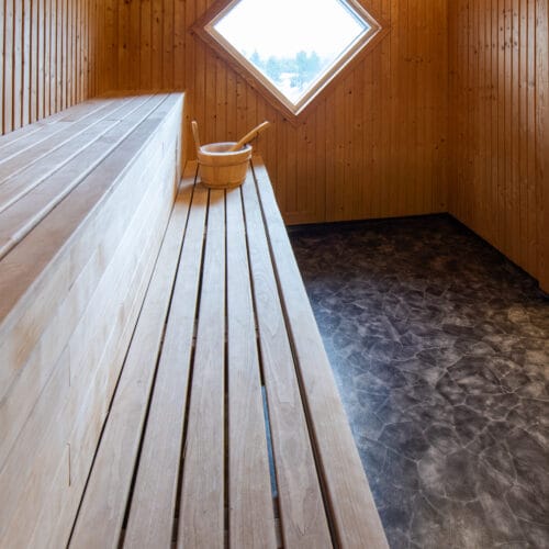 Træbeklædt sauna med træbænke, spand og øse, diamantformet vindue og mørkt gulv.