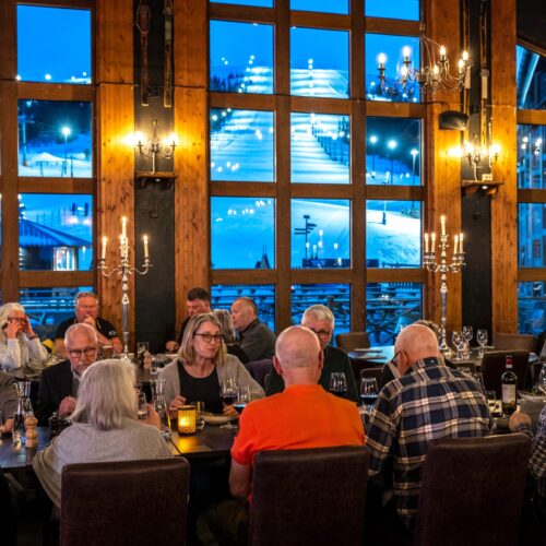 People sitting at long tables in restaurant with large windows, view of illuminated ski slope and snowy landscape outside