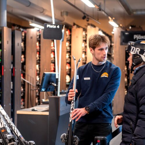 Ski hire where staff show skis to customers at the counter, surrounded by shelves of equipment