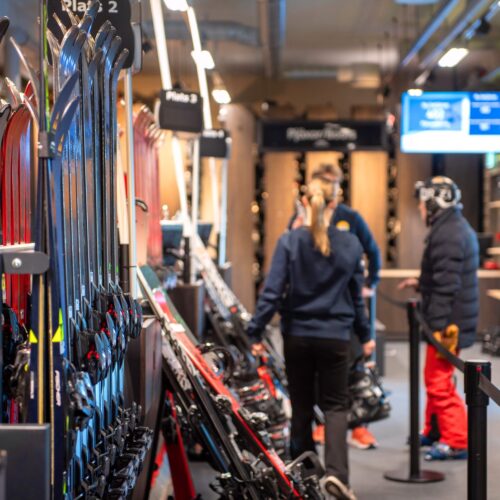 Skiduthyrning med rader av skidor och stavar, personal hjälper kund vid disk i bakgrunden