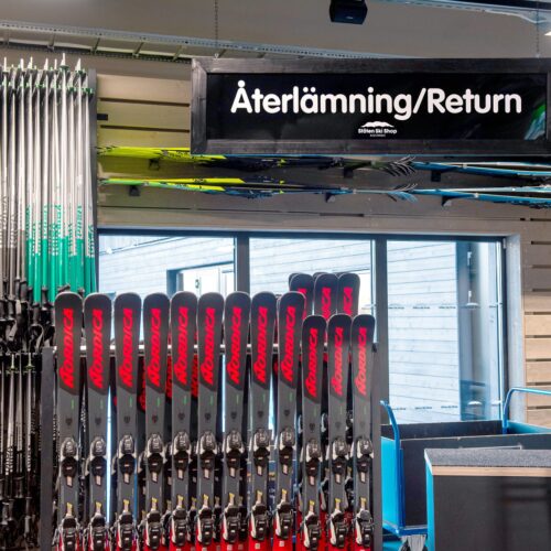 Skiduthyrning med rader av skidor och stavar, skylt för “Återlämning/Return” ovanför disken