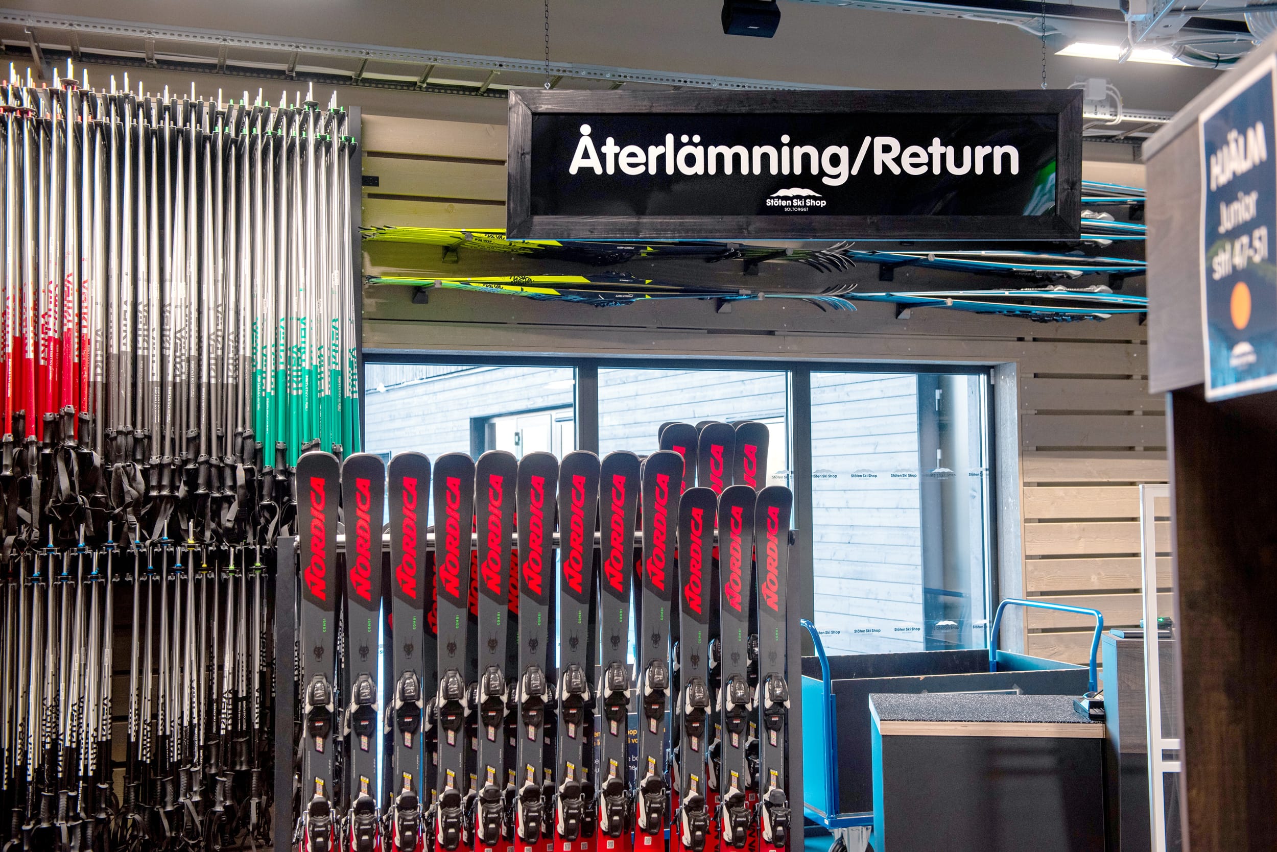 Skiduthyrning med rader av skidor och stavar, skylt för “Återlämning/Return” ovanför disken