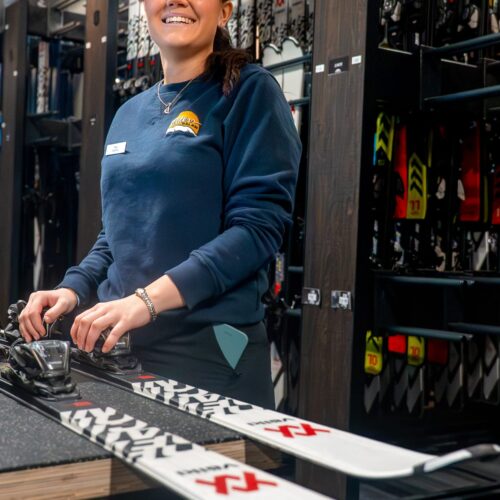 Person justerer skibinding med værktøj i skiudlejningsbutik, omgivet af ski på hylder