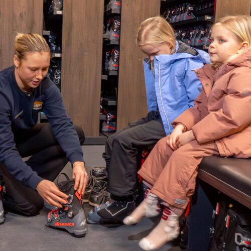 Två barn sitter i skiduthyrning med pjäxor framför sig, personal hjälper till vid provning