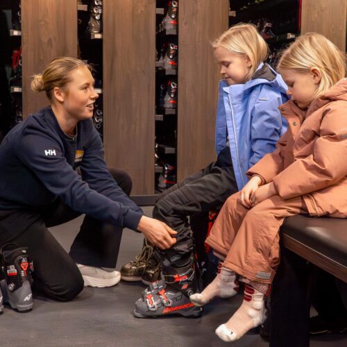 To børn sidder i skileje med skistøvler foran sig, personale hjælper med afprøvning
