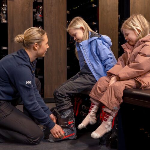 Två barn sitter i skiduthyrning med pjäxor framför sig, personal hjälper till vid provning