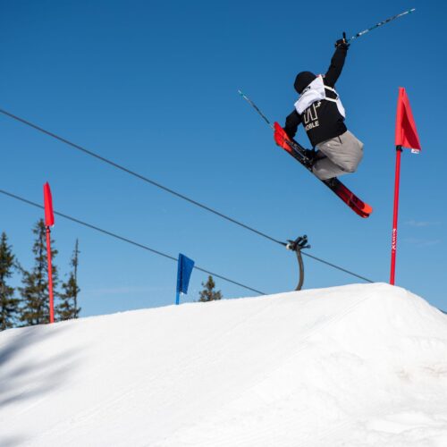 Skiløbere hopper over sneklædte spring med krydsede ski under klar blå himmel og farvede flag