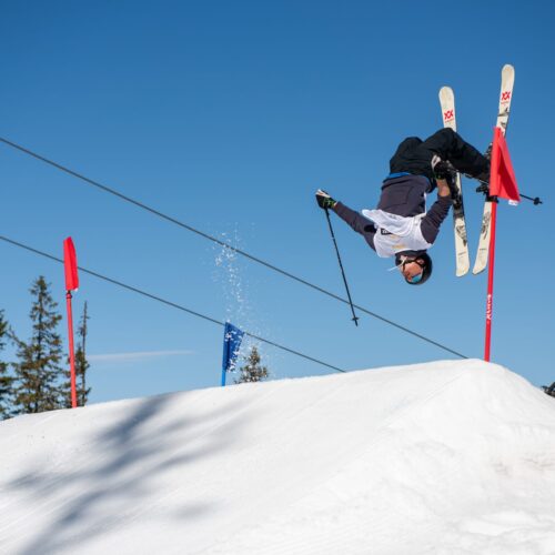 Skiløbere laver baglæns saltomortaler over snedækket hoppekant under klar blå himmel med flag i baggrunden