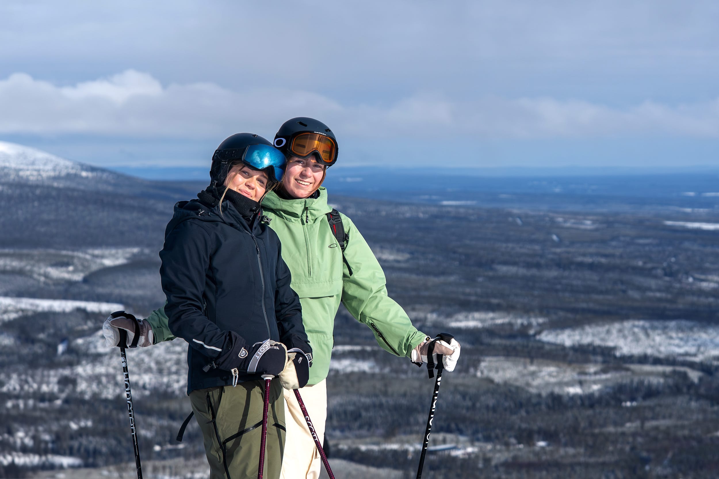 To skiløbere står med stave på et snedækket bjerg, stort landskab og overskyet himmel i baggrunden