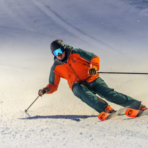 Skier in orange jacket makes sharp turns on groomed snowy ski slope