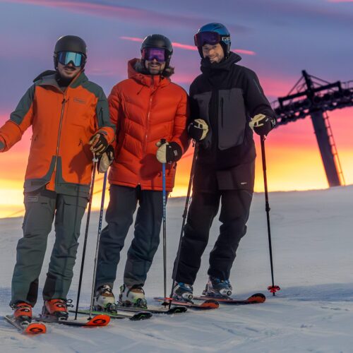 Tre skidåkare står på snötäckt backe vid solnedgång, lift i bakgrunden
