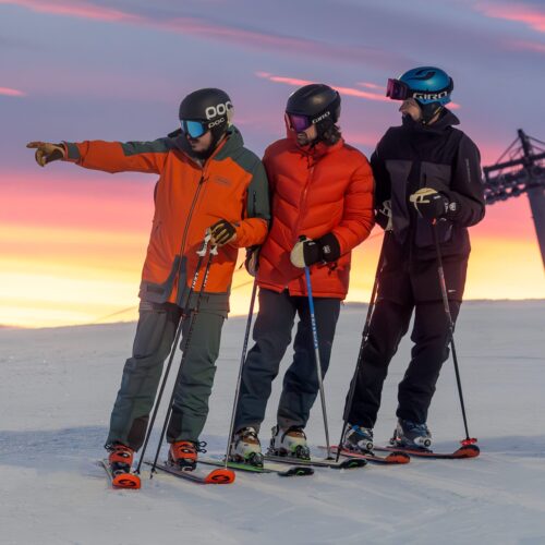Three skiers standing on snow-covered slope at sunset, pointing to mountain and lift